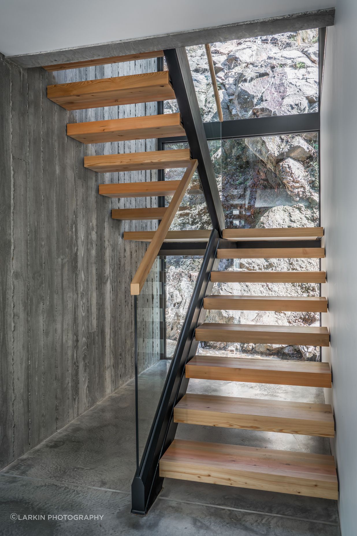Double-height glazing exposes the rugged cliff face upon entry, forming a textural, organic backdrop to the timber staircase.